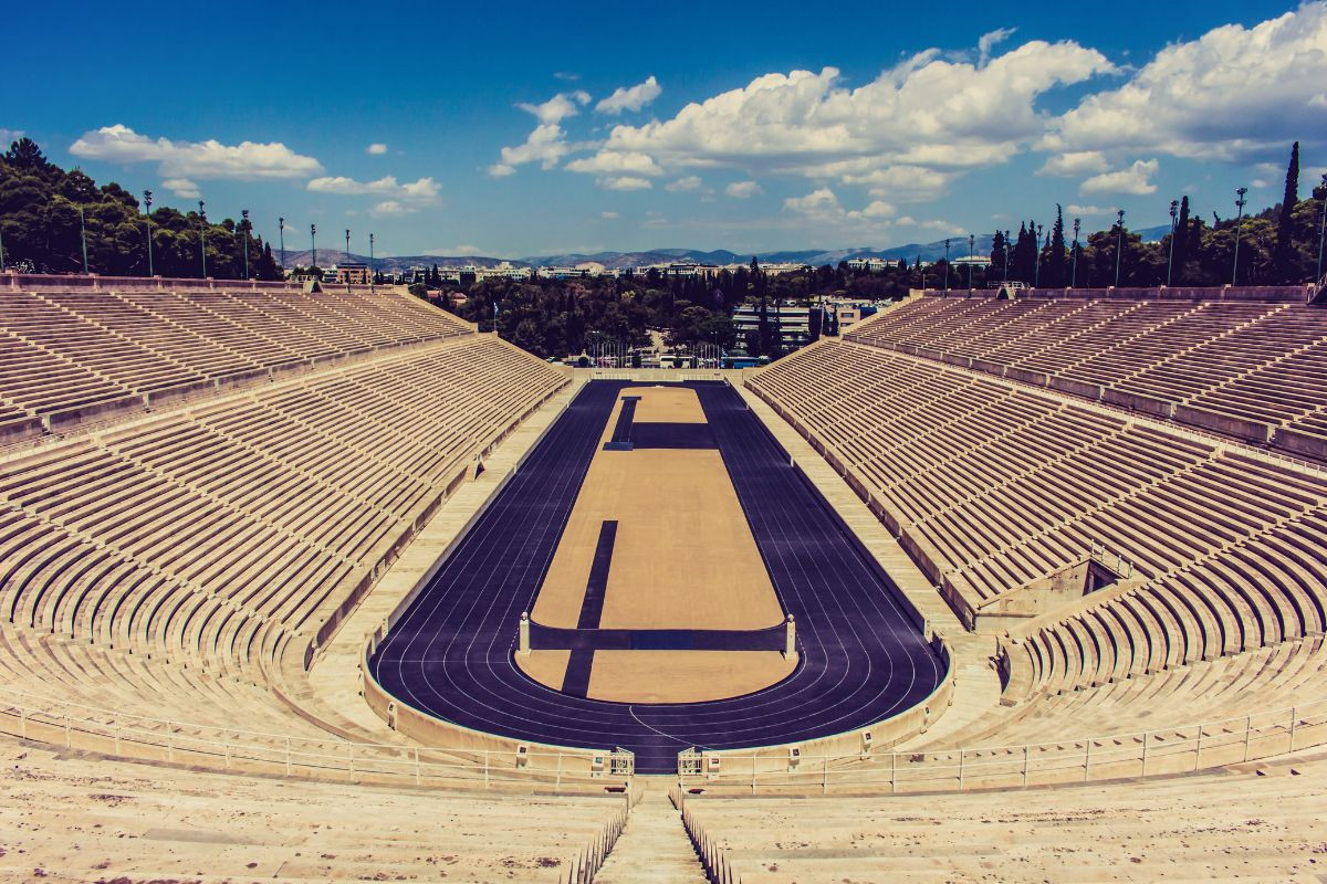 The Panathenaic Stadium in Athens, Greece, an ancient marble stadium with tiered seating and a running track, set against a backdrop of trees and mountains.